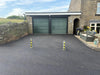 Two Ramco 100 telescopic security bollards with hi-vis tape installed on tarmac driveway in front of stone-built house with double garage