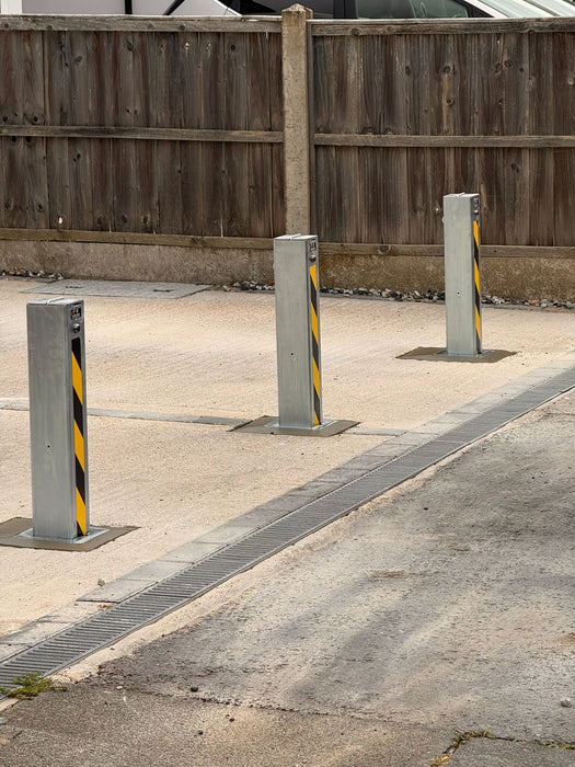 Three Ramco 100 telescopic security bollards with hi-vis chevron tape securing a wide driveway entrance — alternative angle view