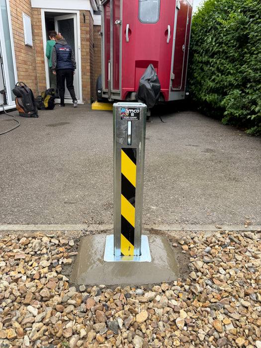 Ramco 100 telescopic security bollard with hi-vis tape freshly installed in concrete on a gravel driveway — close-up of base plate installation